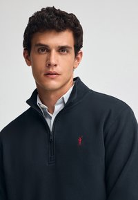 Sudadera de forro polar negra con cremallera y cuello alto, adornada con un logotipo rojo bordado en el pecho, superpuesta sobre una camisa de vestir de rayas azules.