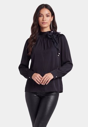 Frau mit langen dunklen Haaren, die eine schwarze Bluse mit langen Ärmeln, Rüschen-Schleife und Perlendetails sowie schwarze Lederhose trägt, steht mit verschränkten Händen.
