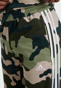 Jogger con stampa mimetica in verde e beige, con un design a tre strisce bianche sul lato e un logo Adidas sulla coscia.
