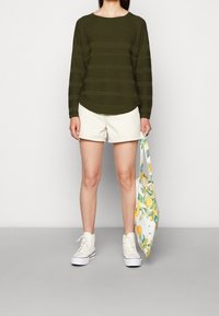 Olivgrünes, strukturiertes Langarmoberteil mit horizontalen Streifen; weiße Shorts; weiße High-Top-Sneaker; trägt eine weiße Tasche mit Zitronenmuster.