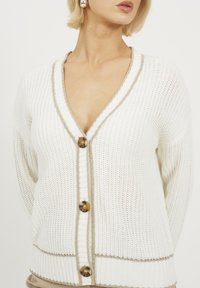 Biały sweter z dzianiny z dekoltem w kształcie litery V, beżowym wykończeniem wzdłuż brzegów i dużymi brązowymi guzikami. Charakteryzuje się luźnym krojem i teksturowanym wzorem.