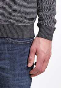 Maglione grigio lavorato a maglia con un motivo testurizzato, polsini a coste e un'etichetta nera, abbinato a jeans in denim blu. In primo piano, una mano che tiene i jeans.