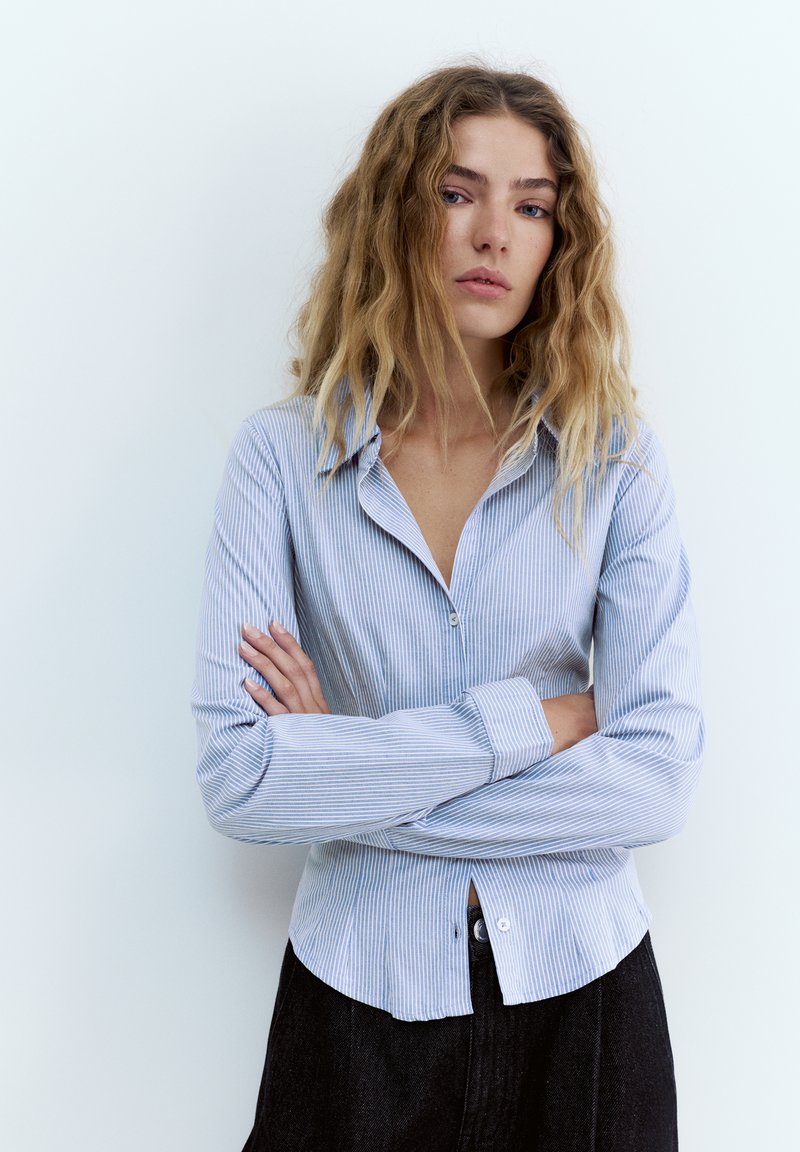 Jeune femme aux cheveux blonds ondulés, portant une chemise à rayures bleu clair boutonnée et un pantalon noir, debout les bras croisés.