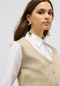 Chaleco de punto beige con escote en forma de V y tres botones, superpuesto a una camisa blanca con botones. La modelo lleva pendientes de oro en forma de gota.