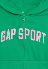 Zelená mikina s kapucňou na zips s veľkým ružovým vyšívaným nápisom "GAP SPORT". Hladký materiál s kapucňou a zipsovým zapínaním vpredu.