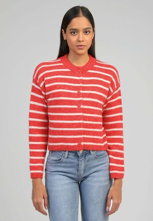 Femme aux longs cheveux bruns portant un cardigan à rayures rouges et blanches boutonné et un jean bleu clair, debout devant un fond uni.