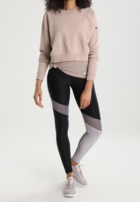 Sweat-shirt court rose clair avec un détail noué, associé à des leggings noirs et gris avec un design en blocs de couleur angulaires. Baskets grises.