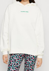 Felpa crema con scritta verde "COLOURFUL REBEL" centrata sul petto. Abbinata a leggings neri con stampa floreale colorata.