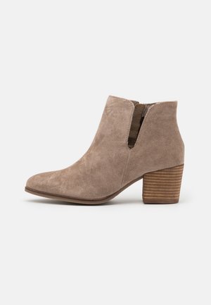 Bottine en daim taupe avec talon carré superposé, panneau élastique latéral et bout arrondi, présentée sur un fond blanc.