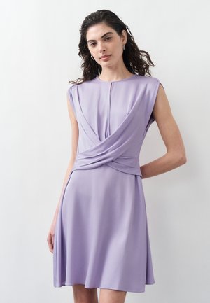 Naine lokkis tume juustega, kes kannab lillakat varrukateta kleiti, millel on vöö juures keerutatud drapeeringu detail, valge taustaga.