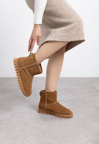 Botas de tobillo de ante marrón con suela de goma gruesa, puntera redondeada y costuras decorativas. El diseño incluye una tira en la parte trasera para facilitar su uso.