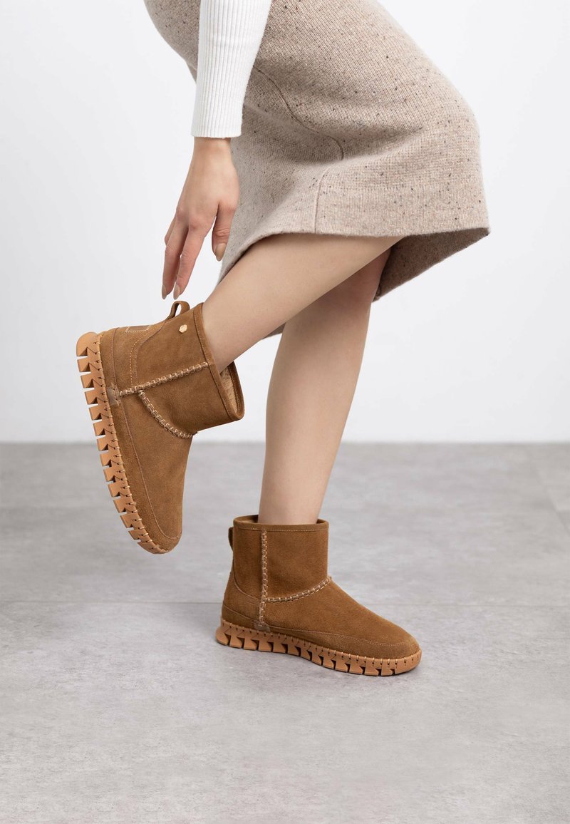 Botas de tobillo de ante marrón con suela de goma gruesa, puntera redondeada y costuras decorativas. El diseño incluye una tira en la parte trasera para facilitar su uso.