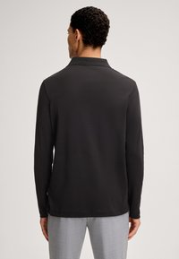 Schwarzes Longsleeve-Polohemd aus glattem Stoff, mit klassischem Kragen und Seitenschlitzen, kombiniert mit hellgrauen Hosen.
