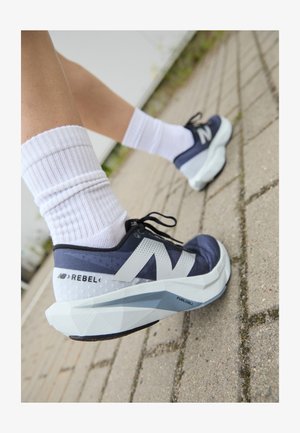 Blaue und weiße Sportschuhe mit einem Obermaterial aus Mesh, einer markanten angularen Zwischensohle und 'REBEL'-Branding. Getragen mit weißen gerippten Socken auf einem befestigten Untergrund.