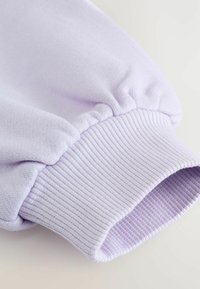 Gros plan sur une manche de sweat-shirt pastel violet avec un tissu froncé et un détail de poignet côtelé sur un fond clair.