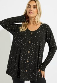 Tunique noire à manches longues avec des pois dorés, texture côtelée, encolure ronde et boutons sur le devant. Silhouette fluide avec une coupe décontractée.