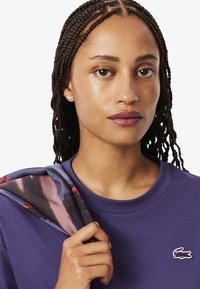 T-shirt viola con scollatura rotonda e logo ricamato. Il modello tiene una sciarpa a fantasia, con tonalità di grigio e rosso, sulla spalla.