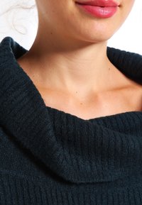 Ciemnozielony sweter w prążki z szerokim dekoltem typu cowl. Tekstura wydaje się miękka, a materiał ma lekki połysk. Kolor skóry widoczny wokół kołnierza.