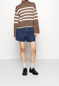 Person i brunt och vitt randigt polokrage, blå denimshorts, vita strumpor och svarta loafer-skor mot vit bakgrund.