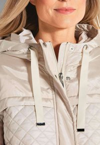 Beige gewatteerde jas met een quilted patroon, zilverkleurige hardware en een capuchon met koordjes. Voorzien van een rits en drukknoopsluiting.