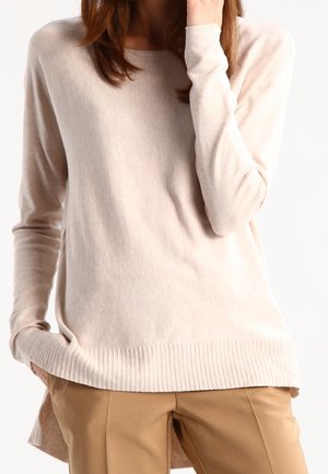 Frau trägt einen hellbeigen Pullover mit langen Ärmeln und camel-farbene Hose, eine Hand in der Tasche und die andere nahe am Gesicht.