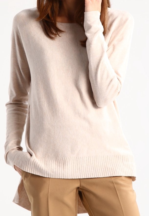 Femme portant un pull beige clair à manches longues et un pantalon couleur camel, une main dans la poche et l'autre près du visage.