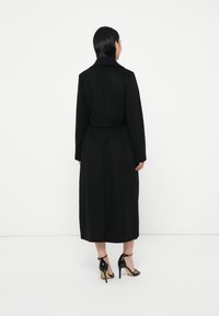 Svart ullkåpe med belte, lange ermer og krage. Den slim-fit designen faller til midten av leggen, sammen med svarte stroppehøye hæler.