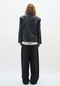 Giubbotto di pelle nera con colletto classico e polsini con cerniera, abbinato a pantaloni neri larghi e una camicia di colore chiaro sotto.
