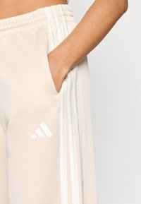 Svetlo bež športne hlače z mehko, gladko teksturo, z belo tridimenzionalno črtastimi poudarki in logotipom Adidas na levi stegni.
