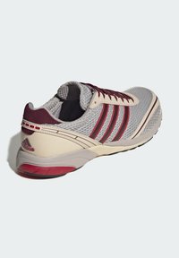 Γκρι και μαρόν αθλητικό παπούτσι Adidas με πλεκτό επάνω μέρος, τρεις ραφές στο πλάι, μαξιλαρωμένη φτέρνα και κόκκινες λεπτομέρειες στα κορδόνια και στην σόλα.