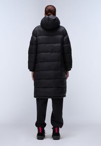 Zwarte pufferjas met een capuchon, gekenmerkt door horizontaal gestikte panelen, lange mouwen en een lengte tot halverwege de kuit. Draagt met zwarte broek en laarzen.