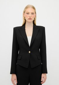 Blazer nero su misura con rever a punta, chiusura con bottone dorato e due tasche frontali. Texture liscia, forma aderente e design minimalista.