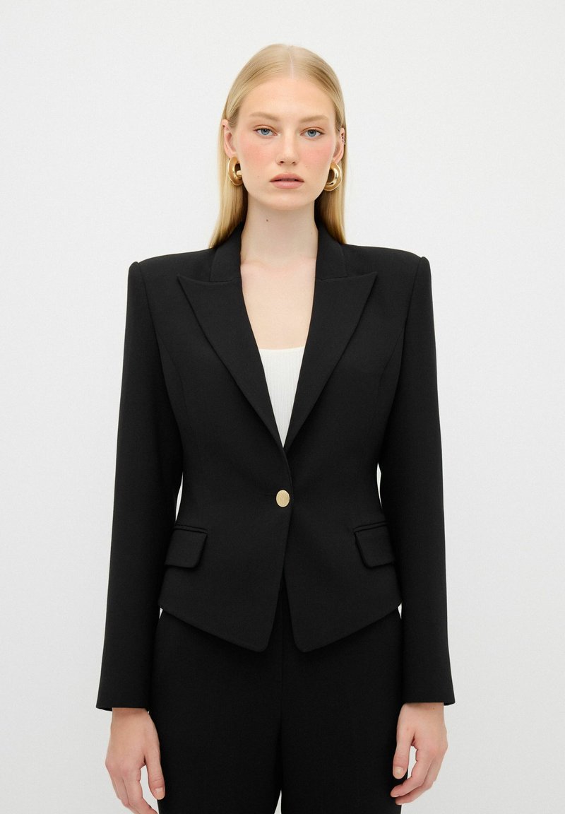 Blazer nero su misura con rever a punta, chiusura con bottone dorato e due tasche frontali. Texture liscia, forma aderente e design minimalista.