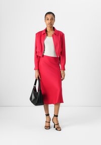 Veste zippée rouge sur un débardeur blanc, associée à une jupe rouge longueur genoux. Un sac à main noir et des talons strapontins complètent la tenue.