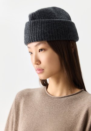 Junge Frau mit glatten dunklen Haaren, die einen beigen Pullover und eine dunkelgraue gestrickte Mütze vor einem einfarbigen Hintergrund trägt.