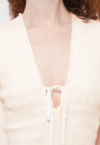 Ελαφρώς μπεζ τοπ με υφασμένο, σφηκοφωλιά σχέδιο. Διαθέτει βαθύ V-λαιμόκοψη και κλείσιμο με κορδόνι μπροστά. Μαλακό, ελαφρύ ύφασμα.