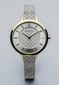 Silver mesh strap watch with a round gold-tone case, textured silver dial, and black hour markers, featuring two black hands and a subtle brand logo.