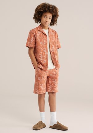 Junge mit lockigem Haar, der ein passendes orangefarbenes Kurzarmhemd mit Blumenmuster und Shorts trägt, weiße Socken und braune Slip-On-Schuhe, Hände in den Taschen.