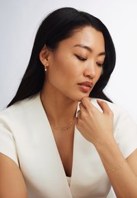 Gold hoop earrings with pearl accents, layered gold chain necklace, and textured gold bracelet. Model wears a white top, showcasing jewelry.