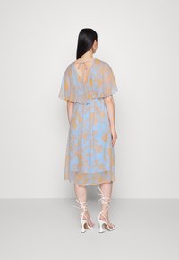 Vestido floral em azul claro com detalhes dourados, mangas curtas e esvoaçantes, costa em forma de chave e uma silhueta leve, combinado com saltos brancos com tiras.