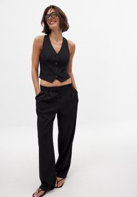 Črn kratki top z petimi gumbi, v kombinaciji s črnimi širokimi hlačami. Gladka tkanina, minimalističen dizajn in usklajeni natikači.