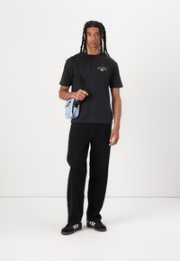 Zwarte T-shirt met grafische print, losse zwarte broek, zwarte sneakers met witte strepen en een kleine lichtblauwe crossbody tas.