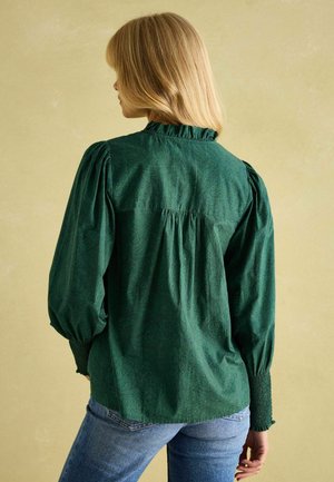 Dunkelgrünbluse mit Paisleymuster, ausgestattet mit einem Rüschensaumkragen, Puffärmeln und gerafftem Rückendetail, kombiniert mit blauen Jeans.