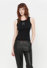 Svart tank topp med en tettsittende design, laget av glatt stoff. Har en liten hvit logo i midten, sammen med høy midje svarte skinnbukser.
