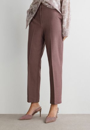 Kobieta ubrana w lawendowe, dopasowane spodnie za kostkę z plisami oraz lawendowe buty na wysokim obcasie z paskiem na pięcie, stojąca na tle gładkiej ściany.