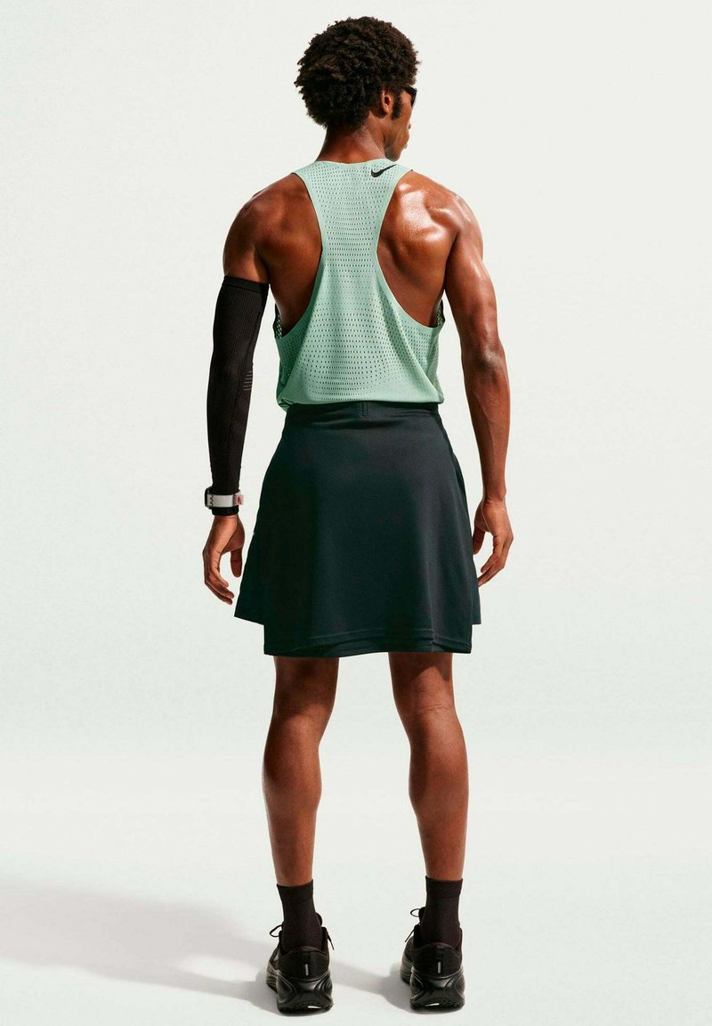 Athlète portant un débardeur menthe, une jupe noire, une manche de compression et des baskets, debout de dos sur un fond uni.