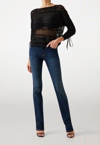 Kobieta nosząca ciemnoniebieskie jeansy o prostych nogawkach, czarną przezroczystą bluzkę na długi rękaw z wzorami oraz czarne buty na obcasie odkryte z przodu, pozująca z jedną ręką w kieszeni.