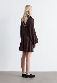 Braunes, strukturiertes Kleid mit langen Ärmeln und einem gemusterten Kragen. Mit schwarzen Slippern getragen, von hinten gezeigt.