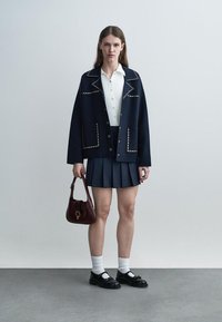 Mladá žena v námornícky modrej bundě s nitmi, biela košeľa na gombíky, námornícky modrá plisovaná sukňa, biele ponožky, čierne topánky, drží bordovú kabelku, stojí na sivej podlahe.