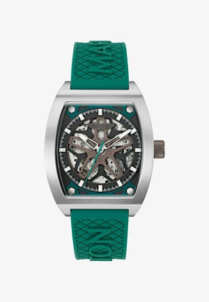 Rustfrit stål armbåndsur med teal gummirem, skeleton urskive der viser indre mekanik, og Aston Martin-logo på skiven.